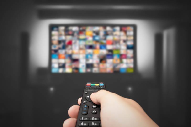 Una mano apunta un control remoto hacia un televisor, en el que se observan cuadritos borrosos simulando diferentes escenas o aplicaciones.