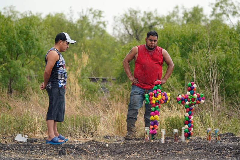 La lluvia cae mientras dos hombres presentan sus respetos en el lugar donde las autoridades encontraron decenas de personas muertas en un semirremolque que contenía presuntos migrantes, el martes 28 de junio de 2022, en San Antonio. (AP Photo/Eric Gay)