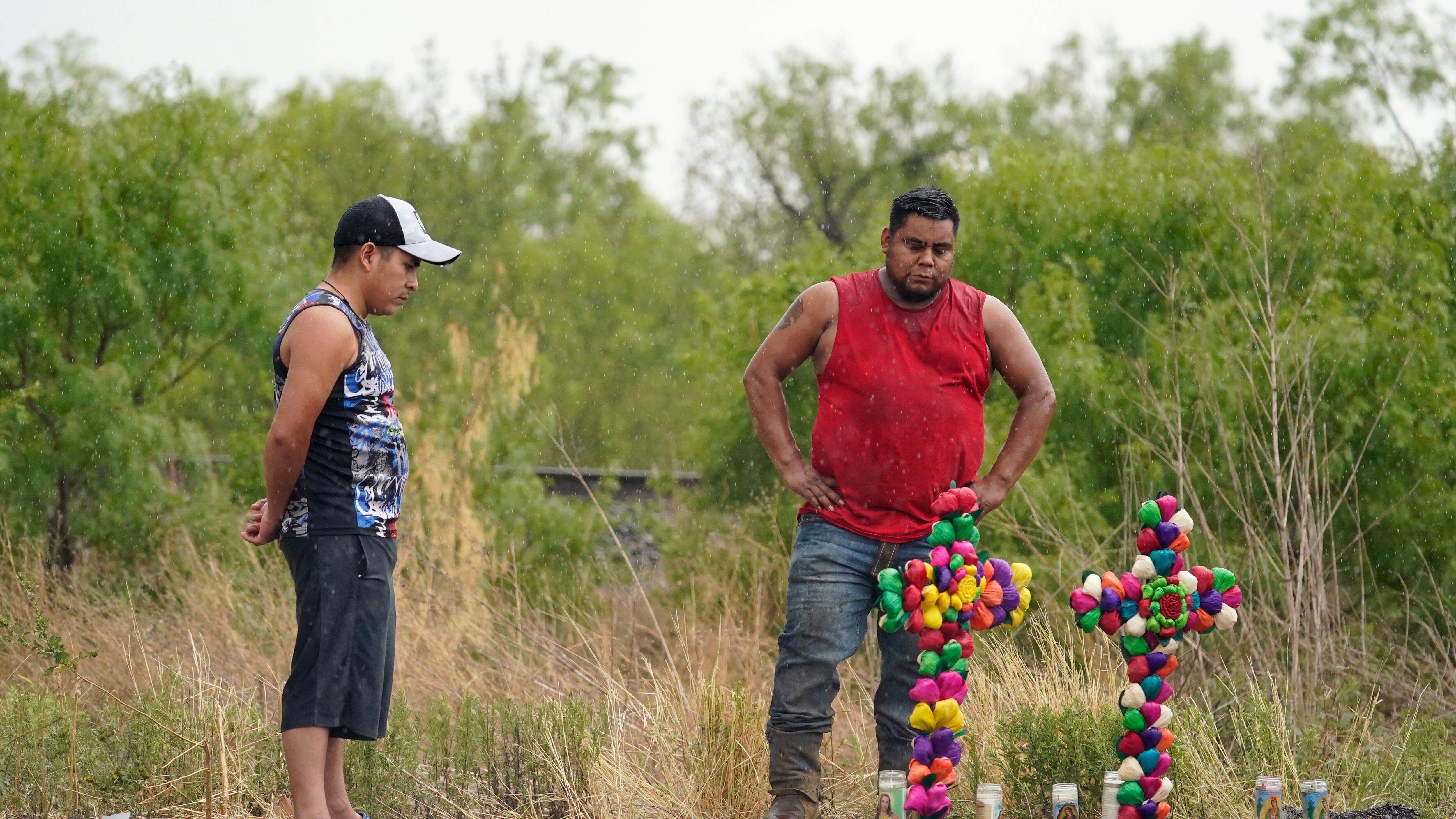 La lluvia cae mientras dos hombres presentan sus respetos en el lugar donde las autoridades encontraron decenas de personas muertas en un semirremolque que contenía presuntos migrantes, el martes 28 de junio de 2022, en San Antonio. (AP Photo/Eric Gay)